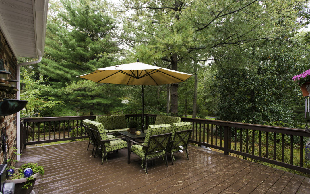 Rain falls on a charming wooden deck with outdoor seating and large yellow umbrella, surrounded by lush greenery for summer-storm-season blog.